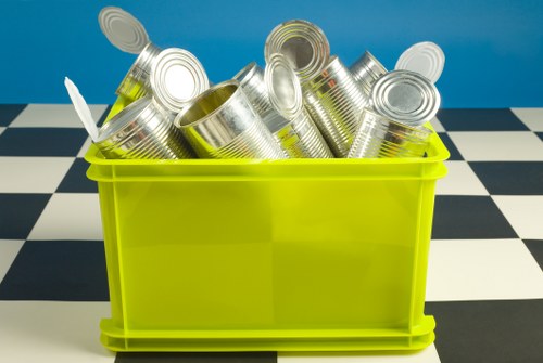A bright yellow plastic container filled with several rolled and crushed aluminium cans, commonly used for beverage containers, placed on a checkered black and white floor. The cans have a shiny metallic surface with visible ridges and are varying in orientation, with some opening upward and others leaning against each other. The background is a plain blue wall, suggesting an indoor setting possibly related to waste collection or recycling. The image illustrates a typical piece of rubbish for removal services, such as those offered by Office Clearance Victoria, highlighting the type of aluminium waste collected during rubbish clearance activities in the local area near Victoria.