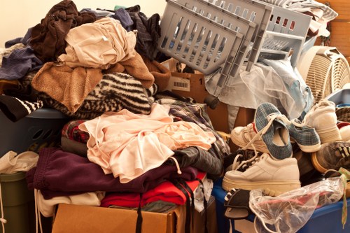 A large pile of mixed household clothing, textiles, and domestic items stacked on top of boxes and a plastic storage container, with some garments including towels, shirts, and sweaters visibly crumpled and overlapping. Among the items are a pair of boxing gloves, a set of sneakers, and various fabric pieces in different colours and textures, including beige, navy, red, and patterned textiles. The collection appears to be situated in an indoor or outdoor space with a wooden surface visible in the background, suggesting a room or storage area prepared for rubbish removal or clearance. The arrangement indicates an ongoing sorting or disposal process, typical of domestic waste clearance services offered by companies like Office Clearance Victoria, located near Victoria, London, serving local household or office cleanouts in the area.