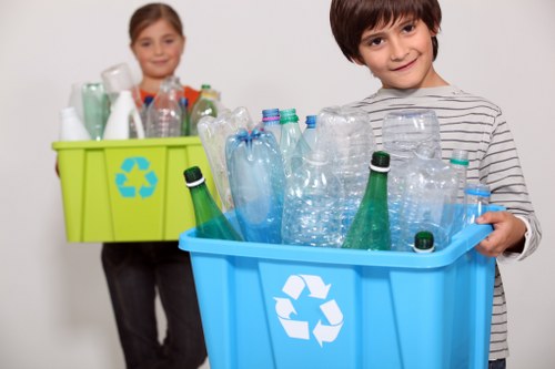 A young boy and girl are standing against a plain white background, each holding a large plastic recycling bin filled with various used plastic bottles. The boy, positioned on the right, has short brown hair and is wearing a grey and white striped long-sleeve shirt, holding a bright blue bin with a white recycling symbol on the front. The bin is filled with clear plastic bottles, some with green and blue caps, and a few bottles are leaning on the sides. The girl, in the background on the left, has her hair tied back and is wearing a grey top with a green recycling bin that features a blue recycling symbol, also filled with empty plastic bottles. Her bin is slightly behind her, and she appears to be in the process of collecting recyclable waste. The scene emphasizes responsible waste sorting and recycling practices, relevant to rubbish removal services in areas like Victoria, UK, with focus on plastic waste management.