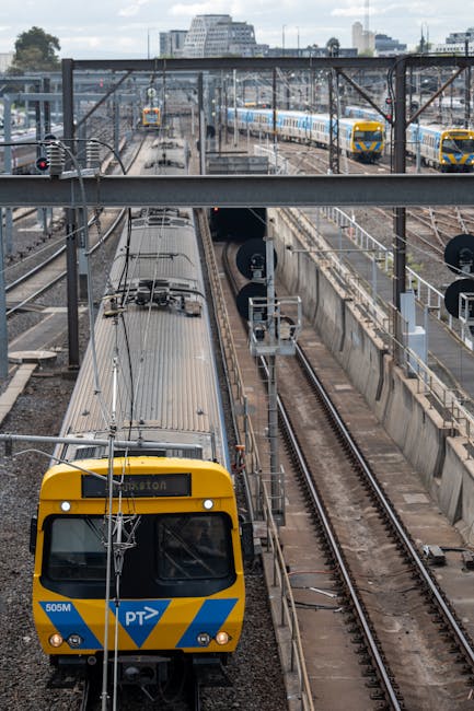 A series of railway tracks extends into the distance within an urban environment, with multiple overhead electric lines and metal supports running parallel above the tracks. To the left, there is a pl