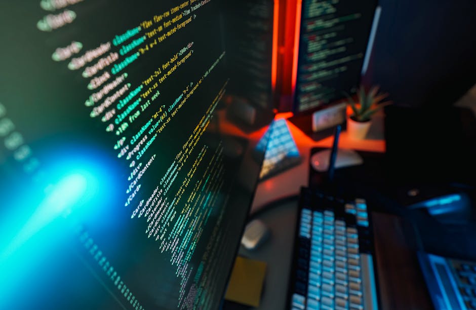 A close-up view of a computer desk featuring a high-resolution monitor displaying colorful programming code with lines of text in shades of white, cyan, and yellow on a dark background. To the right o