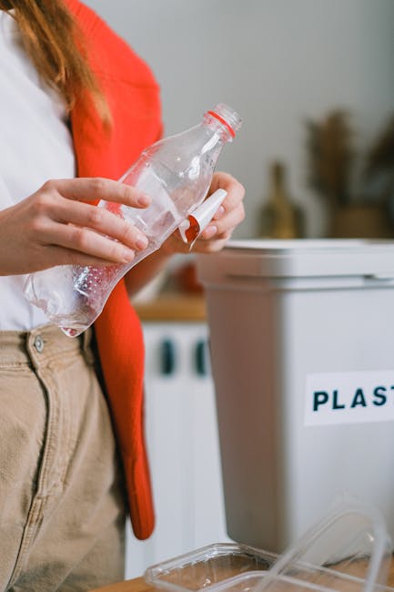 A person wearing orange work overalls and white shoes is standing indoors on a gray carpeted floor, holding two large blue plastic rubbish bags, one in each hand. The bags are filled with waste materi